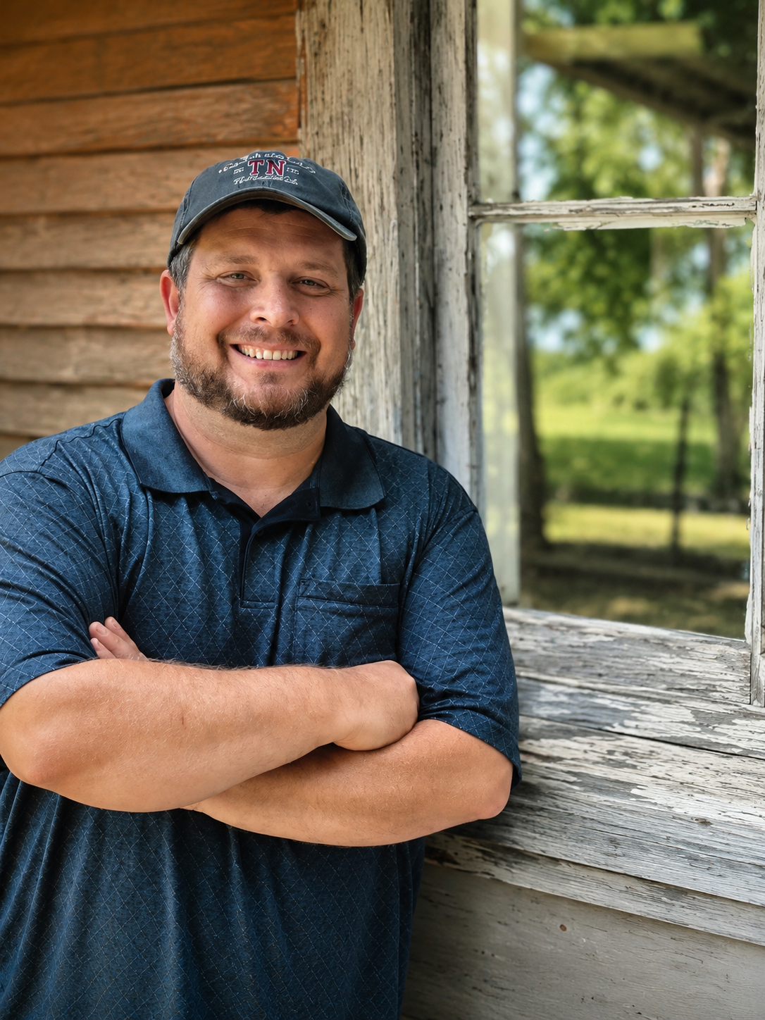 Raymond Alston in a dark polo and cap beside a rustic wooden building